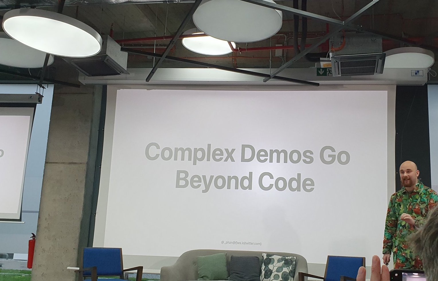 Kevin stands on stage in a forest jumpsuit. His slide reads "Complex Demos Go Beyond Code"